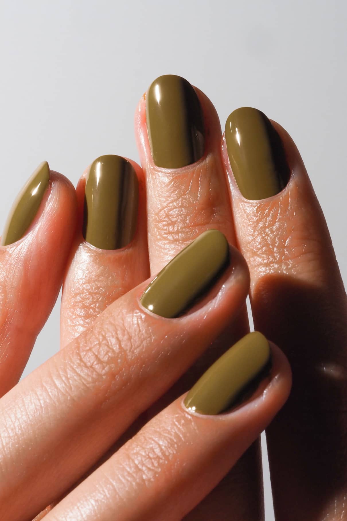 Close-up of a hand with olive green nail polish on a neutral background.